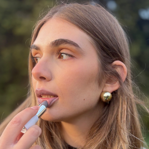 Woman applying vegan natural lipstick outdoors, showcasing soft glam makeup look