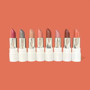 Eight vegan lipsticks in various natural shades with white tubes on a coral background.