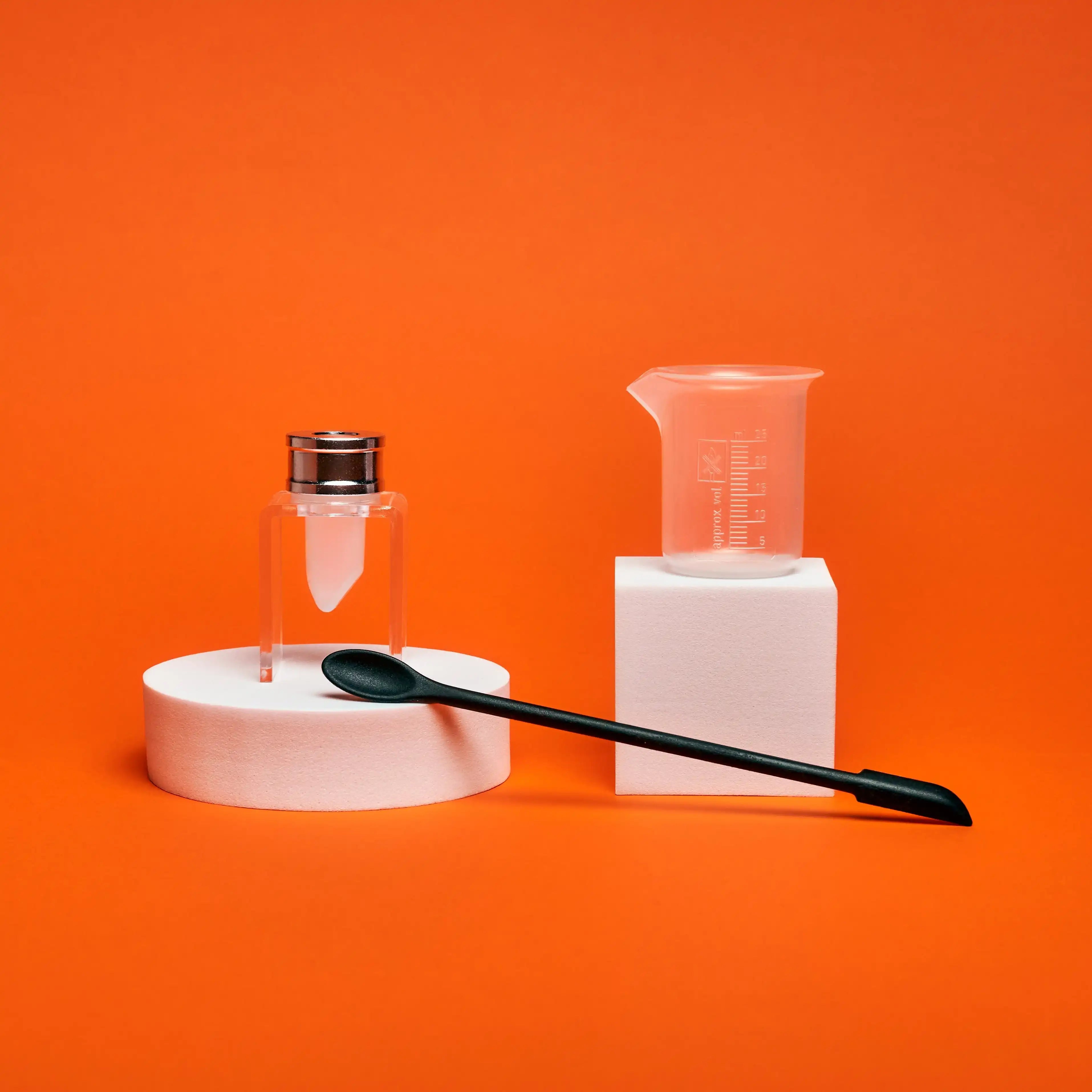DIY cosmetic tools set with glass vial, plastic beaker, and spatula on orange background