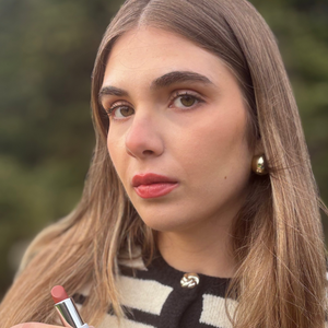 Woman applies natural nude lipstick outdoors, wearing striped top and gold earrings