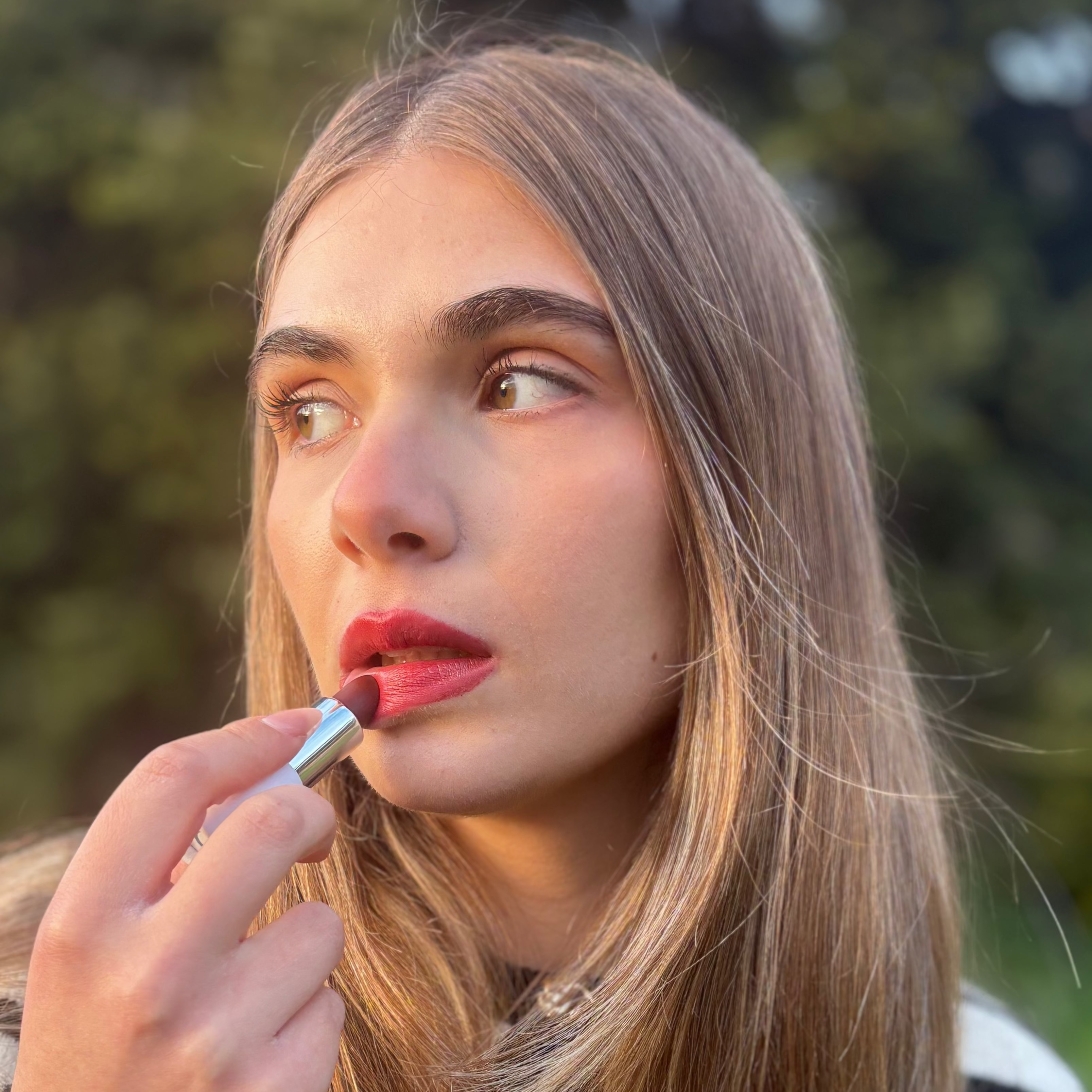 Woman applying natural vegan red lipstick outdoors with sunlight highlighting her face