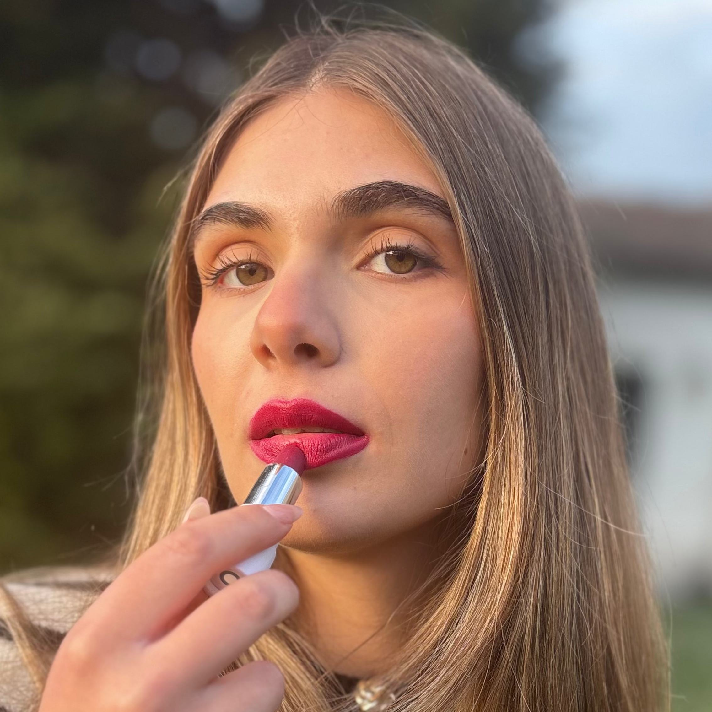 Woman applying vibrant pink vegan lipstick outdoors with natural makeup look
