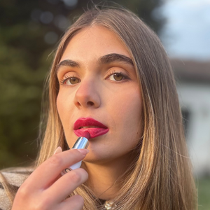 Woman applying vibrant pink vegan lipstick outdoors with natural makeup look